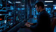In a dimly lit control room, a focused analyst works at his keyboard, illuminated by the glow of intricate digital displays filled with complex data flows, diagrams, and security protocols. The blue-toned interface highlights critical information, reflecting the high-stakes environment of cybersecurity. In the background, colleagues engage in quiet collaboration. Ai generated. Photography style.