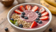 A white bowl filled with creamy pink smoothie is topped with neatly arranged slices of strawberries, plump blueberries, and crunchy granola clusters. A sprig of fresh green basil adds a pop of color, while bananas and additional berries are scattered in the blurred background on a wooden table, creating a wholesome and appetizing scene. Ai generated. Photography style.