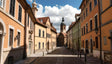 A picturesque narrow street lined with pastel-colored buildings featuring red-tiled roofs and shuttered windows. The cobblestone path leads to a tall church with a dome and a clock tower in the distance. Classic black streetlamps adorn the walls, and the sky above is bright with fluffy white clouds. The scene is serene and charming. Ai generated. Photography style.