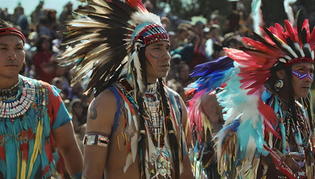 Three Indigenous men wear vibrant ceremonial attire, including large feathered headdresses in shades of red, white, blue, and black. Their regalia features detailed beadwork, painted faces, and traditional accessories. A crowd in the blurred background suggests a public gathering or cultural celebration.