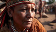 A close-up of an elder Indigenous woman with weathered skin and deep expression lines, wearing a red and green beaded headband adorned with pearls and feathers. Her earrings are handcrafted, featuring natural materials. She wears a necklace made of white and red beads. In the softly blurred background, people are seen standing in a sunlit outdoor gathering.
