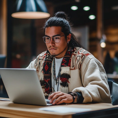 A young man with long dark hair tied back works on a laptop at a wooden table. He wears round glasses, layered necklaces, and a cream-colored jacket adorned with Indigenous patterns in red, black, and beige. The background shows a warmly lit indoor setting with blurred lights and modern furnishings, creating a cozy and studious atmosphere.