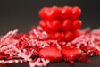 A red heart-shaped candle sits in the background against a dark surface, slightly out of focus. In the foreground, two heart-shaped chocolates wrapped in shiny red foil rest among red and pink crinkled paper shreds. The glossy textures of the candle and chocolates reflect soft light, contrasting with the matte paper decorations. The composition highlights the rich red tones and smooth surfaces. Photography.