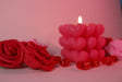 A softly glowing heart-shaped candle stands in the background, slightly out of focus, casting warm light. Two red roses with delicate petals are in the foreground, crisp and detailed. Small red glass hearts are scattered around, reflecting the soft glow. The background blends into a dreamy blur of pink and red tones, enhancing the romantic and gentle feel of the composition. Photography.