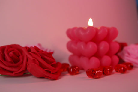 A softly glowing heart-shaped candle stands in the background, slightly out of focus, casting warm light. Two red roses with delicate petals are in the foreground, crisp and detailed. Small red glass hearts are scattered around, reflecting the soft glow. The background blends into a dreamy blur of pink and red tones, enhancing the romantic and gentle feel of the composition. Photography.