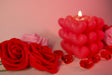 A softly glowing heart-shaped candle stands in the background, slightly out of focus, casting warm light. Two red roses with delicate petals are in the foreground, crisp and detailed. Small red glass hearts are scattered around, reflecting the soft glow. The background blends into a dreamy blur of pink and red tones, enhancing the romantic and gentle feel of the composition. Photography.