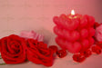 A softly glowing heart-shaped candle stands in the background, slightly out of focus, casting warm light. Two red roses with delicate petals are in the foreground, crisp and detailed. Small red glass hearts are scattered around, reflecting the soft glow. The background blends into a dreamy blur of pink and red tones, enhancing the romantic and gentle feel of the composition. Photography.