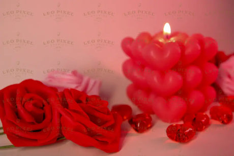 A softly glowing heart-shaped candle stands in the background, slightly out of focus, casting warm light. Two red roses with delicate petals are in the foreground, crisp and detailed. Small red glass hearts are scattered around, reflecting the soft glow. The background blends into a dreamy blur of pink and red tones, enhancing the romantic and gentle feel of the composition. Photography.