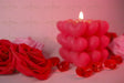 A softly glowing heart-shaped candle stands in the background, slightly out of focus, casting warm light. Two red roses with delicate petals are in the foreground, crisp and detailed. Small red glass hearts are scattered around, reflecting the soft glow. The background blends into a dreamy blur of pink and red tones, enhancing the romantic and gentle feel of the composition. Photography.