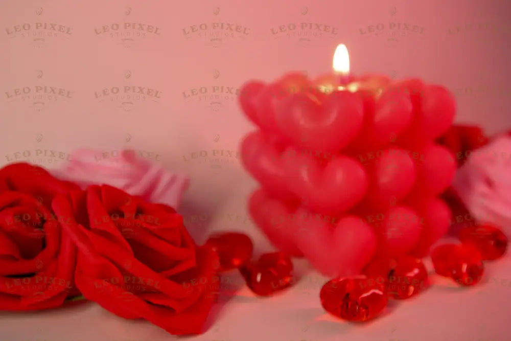 A softly glowing heart-shaped candle stands in the background, slightly out of focus, casting warm light. Two red roses with delicate petals are in the foreground, crisp and detailed. Small red glass hearts are scattered around, reflecting the soft glow. The background blends into a dreamy blur of pink and red tones, enhancing the romantic and gentle feel of the composition. Photography.