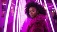 A striking woman is captured in a neon-lit hallway dominated by radiant purple lights. She wears a vibrant purple fur coat, with her natural afro framing her face elegantly. Hoop earrings accentuate her poised expression, and soft bokeh lights in the background add depth to the composition. The glowing vertical neon lights create a futuristic and captivating atmosphere. Ai generated. Photography style.