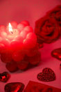 A close-up of a lit red candle shaped like a cluster of small hearts. The candle emits a soft glow, illuminating the smooth wax texture. Surrounding it are deep red artificial roses, glossy heart-shaped decorations, and a glittery heart ornament. The blurred background enhances depth, with the candle as the focal point. The warm lighting and rich tones create an intimate and moody photography style.