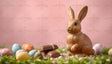 A shiny chocolate bunny figurine is positioned on vibrant green grass, surrounded by colorful eggs in pastel hues of blue, pink, yellow, and orange with dotted designs. Nearby, small chocolate pieces lie on a scattered bed of crumbs. The background is a soft pink gradient, highlighting the festive and playful Easter theme. Ai generated. Photography style.