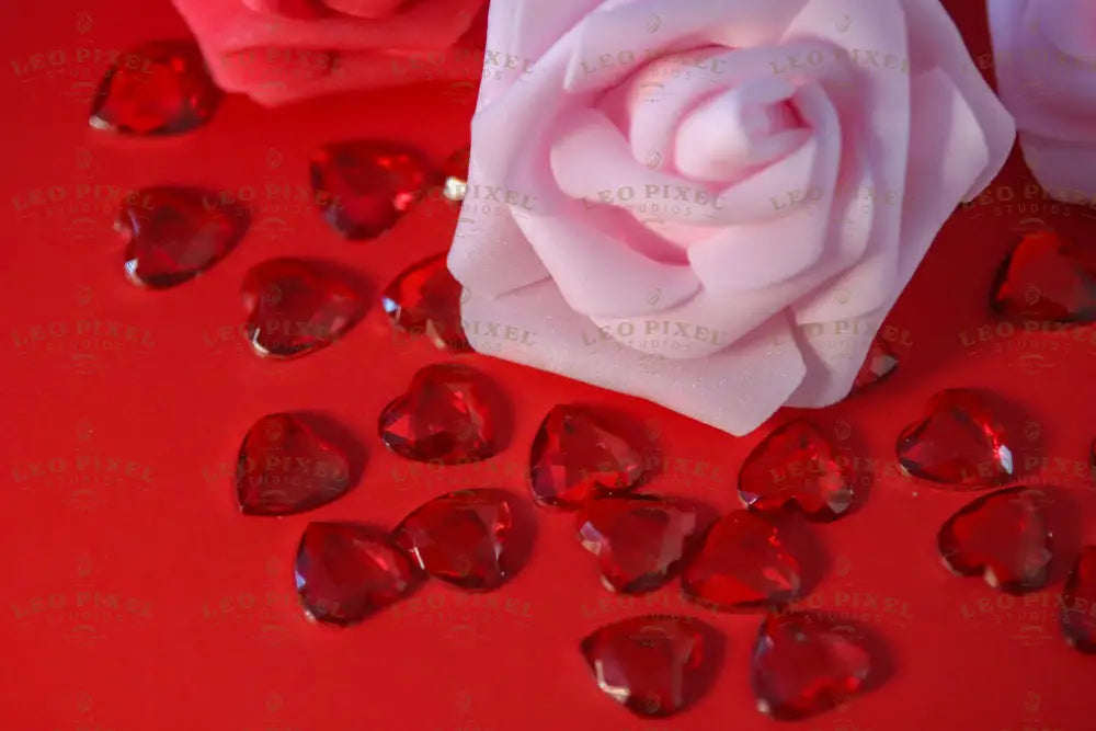 A soft pink rose with smooth, curved petals is at the center, surrounded by scattered red, faceted heart-shaped gems on a red surface. A deeper red rose is slightly blurred in the background, adding depth. The scene is evenly lit, emphasizing the velvety texture of the flowers and the sparkle of the gems. The composition highlights the contrast between the soft petals and the glossy jewels. Photography.