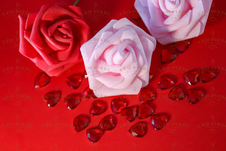 A soft pink artificial rose with delicate, sculpted petals sits in the foreground, surrounded by scattered red, faceted heart-shaped gems on a vibrant red background. A deep red rose and another pale pink rose are positioned slightly behind, adding depth. The reflective gems contrast with the smooth texture of the flowers, and the balanced lighting enhances the colors and details. Photography.