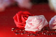 A close-up of a delicate pink rose with smooth, curled petals resting on a red surface. Behind and next to it, a slightly blurred red rose and pink rose adds depth. Scattered around are small, faceted red heart-shaped gems reflecting light. The background has a warm, softly blurred effect, enhancing the romantic tones of the composition. The rich reds and soft pink hues contrast beautifully in this detailed shot.   Photography.