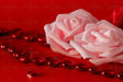 A close-up of two soft pink roses resting on a red surface, their delicate petals curling with a velvety texture. A strand of glossy red heart-shaped beads stretches across, reflecting light with a deep shine. Scattered heart gems add detail, their facets catching subtle highlights. The rich contrast between the pale roses and vibrant red elements creates a visually striking composition. Photography.