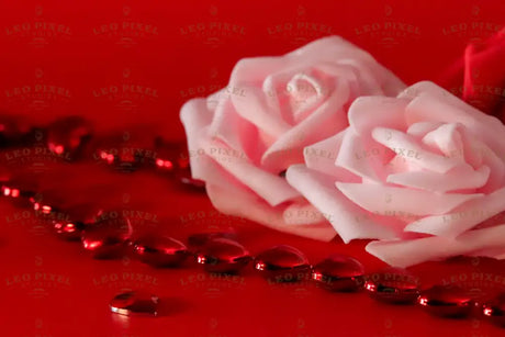 A close-up of two soft pink roses resting on a red surface, their delicate petals curling with a velvety texture. A strand of glossy red heart-shaped beads stretches across, reflecting light with a deep shine. Scattered heart gems add detail, their facets catching subtle highlights. The rich contrast between the pale roses and vibrant red elements creates a visually striking composition. Photography.