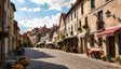 This image shows a charming cobblestone street lined with pastel-colored buildings, adorned with potted plants and flower boxes on balconies. Red and white awnings shade small café tables, inviting visitors to enjoy the setting. The warm sunlight highlights the texture of the walls and rooftops, while a clear blue sky with fluffy clouds enhances the serene atmosphere. Ai generated. Photography style.