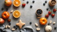 A flat lay composition of fresh ingredients on a smooth gray surface. Sliced oranges, bright red strawberries, and deep blue blueberries are scattered among round tomatoes and a white pumpkin. Small wooden bowls hold nuts and more berries. A soft gray fabric adds texture in the corner. A star-shaped biscuit sits at the center, surrounded by shelled nuts and fresh produce, creating a balanced visual. Ai generated. Photography style.