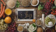 The image shows an assortment of fresh and dry foods spread across a wooden surface. Carrots, orange juice, basil leaves, and grapes surround a small black chalkboard. Bowls hold sunflower seeds, powder, and sliced zucchini. White beans, green grapes, and cherry tomatoes add vibrant color. The arrangement highlights natural foods with textures and colors carefully displayed. Ai generated. Photography style.