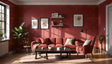 This warm living room features a rich red wall adorned with framed botanical art and wooden floating shelves holding books and decor. A comfortable red sectional sofa and matching armchair are paired with a dark wood coffee table. A plush pink rug, potted greenery, and a black floor lamp add charm. Natural light pours in through large windows, enhancing the cozy and inviting atmosphere. Ai generated. Photography style.