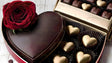  A luxurious heart-shaped chocolate box takes center stage, topped with a velvety red rose, symbolizing deep affection. Nestled beside it are exquisite gold-wrapped heart chocolates, each gleaming in black paper cups. A golden box filled with assorted treats peeks in the background, completing this romantic, indulgent setting on a softly lit wooden surface. Ai generated image.