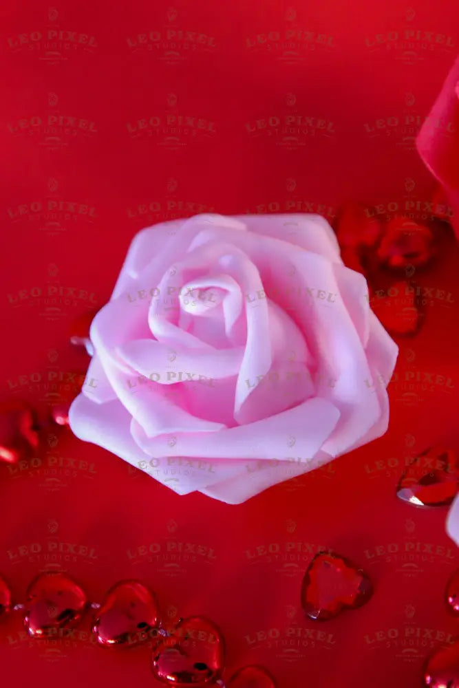 A soft pink artificial rose rests on a red surface, surrounded by shiny red heart-shaped beads and scattered gems. The petals have a delicate texture, contrasting with the reflective surfaces of the hearts. The background remains vibrant red, complementing the rich tones of the decorative elements. The lighting enhances the depth and smooth texture of the petals and the glossy finish of the beads. Photography.