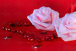 A soft pink rose rests on a red surface, accompanied by two additional roses—one red and another soft pink, slightly out of focus. Surrounding them are shiny red heart-shaped beads and scattered gems. The petals have a smooth texture, contrasting with the reflective surfaces of the hearts. The rich red background enhances the depth, highlighting the delicate petals and glossy beads. Photography.