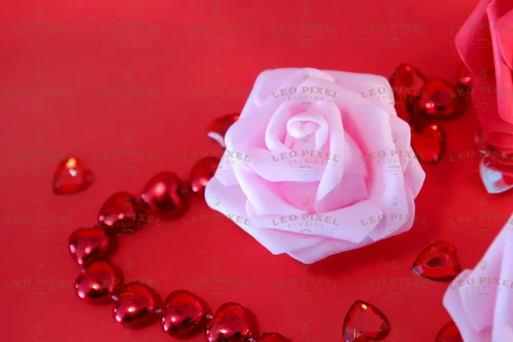 A soft pink rose rests on a red surface, accompanied by two additional roses—one red and another soft pink, slightly out of focus. Surrounding them are shiny red heart-shaped beads and scattered gems. The petals have a smooth texture, contrasting with the reflective surfaces of the hearts. The rich red background enhances the depth, highlighting the delicate petals and glossy beads. Photography.