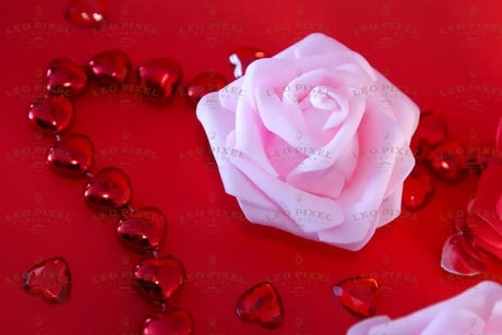 A soft pink rose rests on a red surface, accompanied by two additional roses—one red and another soft pink, slightly out of focus. Surrounding them are shiny red heart-shaped beads and scattered gems. The petals have a smooth texture, contrasting with the reflective surfaces of the hearts. The rich red background enhances the depth, highlighting the delicate petals and glossy beads. Photography.