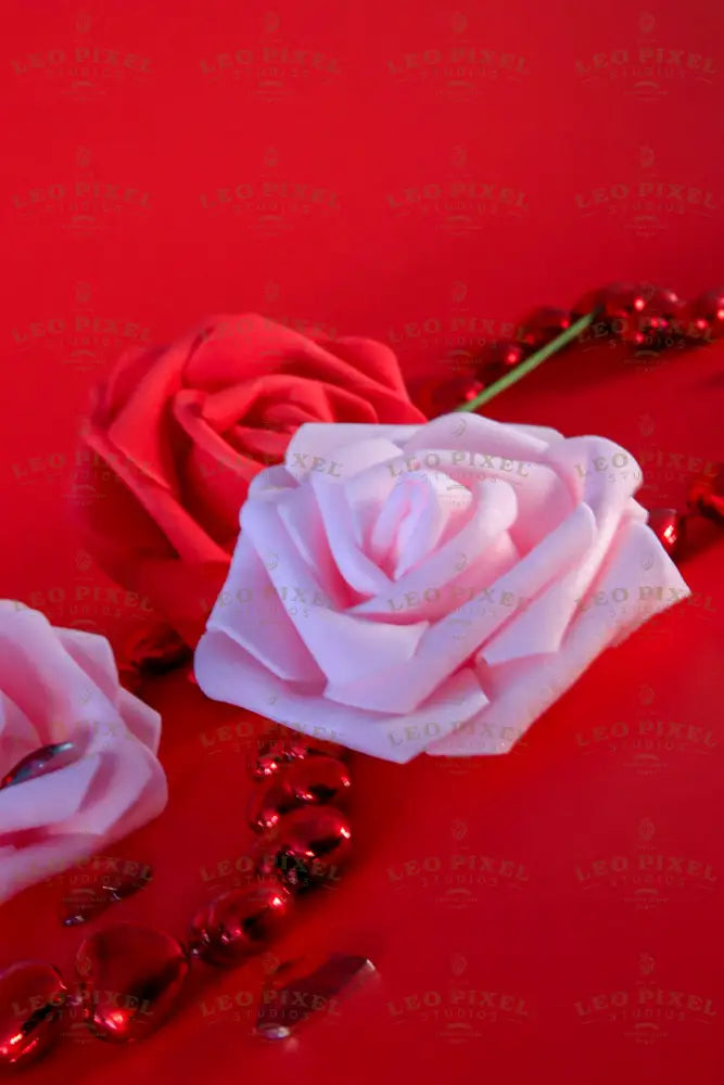 A soft pink artificial rose sits in focus against a red background, with a red rose behind it and another pink rose slightly visible to the side. A string of glossy red heart-shaped beads winds around them, reflecting light. Scattered red heart gems add texture. The smooth petals contrast with the shiny decorations, creating a visually rich composition in a close-up photography style. 