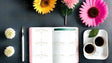 Flat lay image of an open notebook with a pink edge and a green header on a dark surface. To the left, two small white flowers and a white pen with a gold tip are neatly arranged. Above the notebook, yellow, pink, and bright pink daisy-like flowers add color. On the right, two white cups filled with black coffee rest on a white tray with a green leaf. The composition is clean and organized, with balanced elements. Ai generated. Photography style.