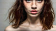 A close-up portrait of a young woman with striking blue-green eyes, scattered freckles across her pale complexion, and softly parted lips in a natural pink hue. Her shoulder-length, tousled brown hair flows loosely, framing her face and neck. The soft lighting enhances the natural texture of her skin and the depth of her gaze, set against a plain, light background. Ai generated. Photography style.