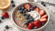 A white bowl holds a creamy yogurt base with a swirl of whipped yogurt on top. Sliced strawberries, whole blueberries, and a layer of granola decorate the bowl. The colors are vibrant, with red, blue, and beige blending beautifully. The background features a lemon half, scattered blueberries, and a silver spoon, all on a smooth white surface, adding a clean and fresh look. Ai generated. Photography style.