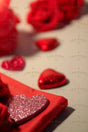 A close-up photograph shows a sparkling pink heart-shaped decoration resting on a neatly folded red fabric. In the background, red foil-wrapped heart chocolates and artificial red roses are softly blurred. The smooth surface reflects the warm tones, while the shallow depth of field draws attention to the glittering heart, creating a soft, romantic mood with rich red hues.