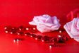 A soft pink rose lies on a red surface, slightly out of focus, accompanied by two other roses—one pink and one red. A string of shiny red heart-shaped beads stretches across the frame, reflecting light. Scattered glossy heart gems add texture. The red background enhances the contrast, emphasizing the smooth petals and reflective details in a close-up photography style. 