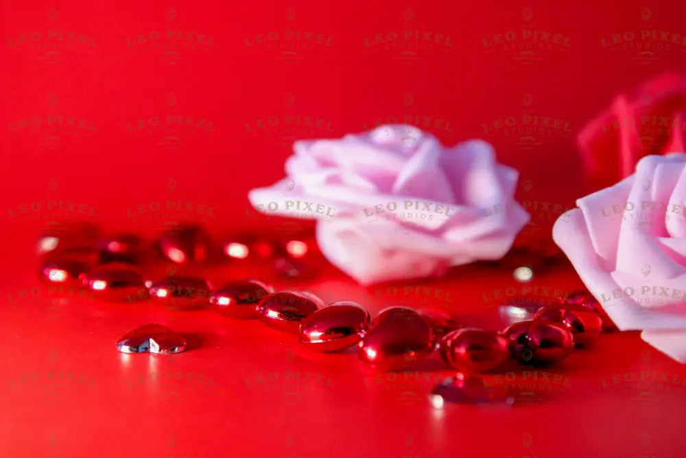 A soft pink rose lies on a red surface, slightly out of focus, accompanied by two other roses—one pink and one red. A string of shiny red heart-shaped beads stretches across the frame, reflecting light. Scattered glossy heart gems add texture. The red background enhances the contrast, emphasizing the smooth petals and reflective details in a close-up photography style. 