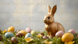 The image features a shiny golden bunny figurine sitting on vibrant green grass. Surrounding it are pastel-colored eggs in shades of blue, yellow, pink, and brown, some with dotted designs. Yellow flowers are scattered among the eggs, adding brightness. The background is softly blurred in neutral tones, enhancing the focus on the bunny and eggs. Ai generated. Photography style.