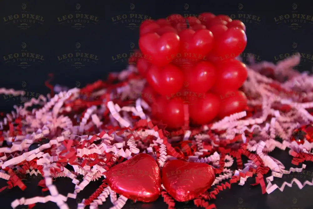 A red heart-shaped candle sits in the background against a dark surface, slightly out of focus. In the foreground, two heart-shaped chocolates wrapped in shiny red foil rest among red and pink crinkled paper shreds. The glossy textures of the candle and chocolates reflect soft light, contrasting with the matte paper decorations. The composition highlights the rich red tones and smooth surfaces. Photography.