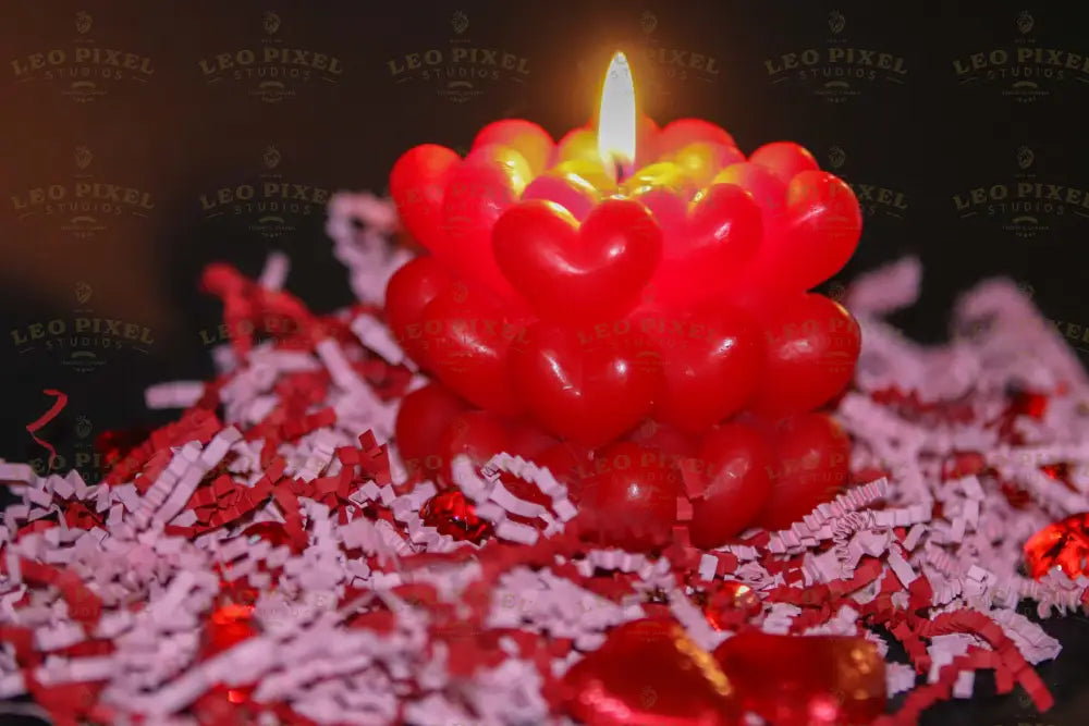 A red candle with heart-shaped wax layers is lit against a dark background. The warm flame casts a soft glow, highlighting the glossy texture of the hearts. The lower part of the image shows scattered decorative elements in shades of red and white, slightly out of focus. The contrast between the candle’s vibrant red and the deep shadows creates an intimate and cozy atmosphere. Photography.