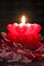 A red candle with heart-shaped wax layers is lit against a dark background. The warm flame casts a soft glow, highlighting the glossy texture of the hearts. The lower part of the image shows scattered decorative elements in shades of red and white, slightly out of focus. The contrast between the candle’s vibrant red and the deep shadows creates an intimate and cozy atmosphere. Photography.