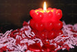 A red candle with heart-shaped wax layers is lit against a dark background. The warm flame casts a soft glow, highlighting the glossy texture of the hearts. The lower part of the image shows scattered decorative elements in shades of red and white, slightly out of focus. The contrast between the candle’s vibrant red and the deep shadows creates an intimate and cozy atmosphere. Photography.