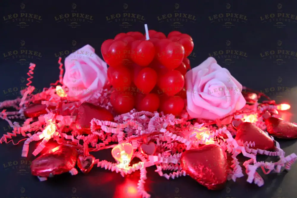 A red heart-shaped cube candle sits in the center, unlit, surrounded by two soft pink foam roses. Red foil-wrapped chocolates and crinkled pink paper fill the foreground. Warm heart-shaped string lights illuminate the scene against a dark background. The deep reds and soft pinks contrast beautifully, with the reflections adding depth and warmth to the composition. Photography.