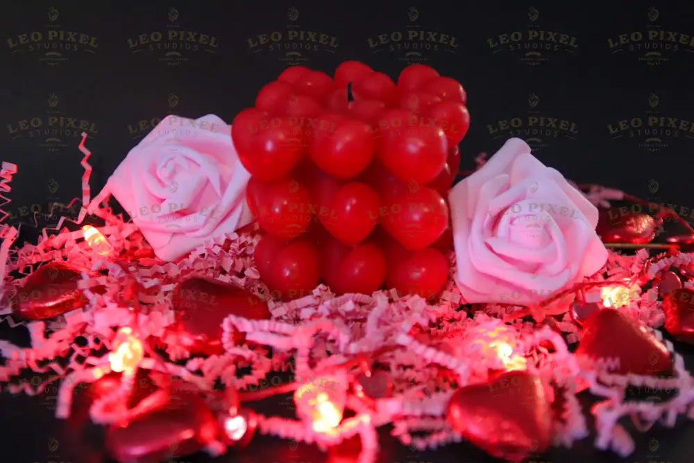 A red heart-shaped cube candle sits in the center, surrounded by pink foam roses and glowing red string lights. The candle’s glossy hearts reflect the warm lighting. Shiny red foil-wrapped chocolates and soft crinkled paper shreds add texture to the dark background. The soft glow of the lights enhances the contrast between the deep reds and delicate pinks, creating a balanced composition. Photography.