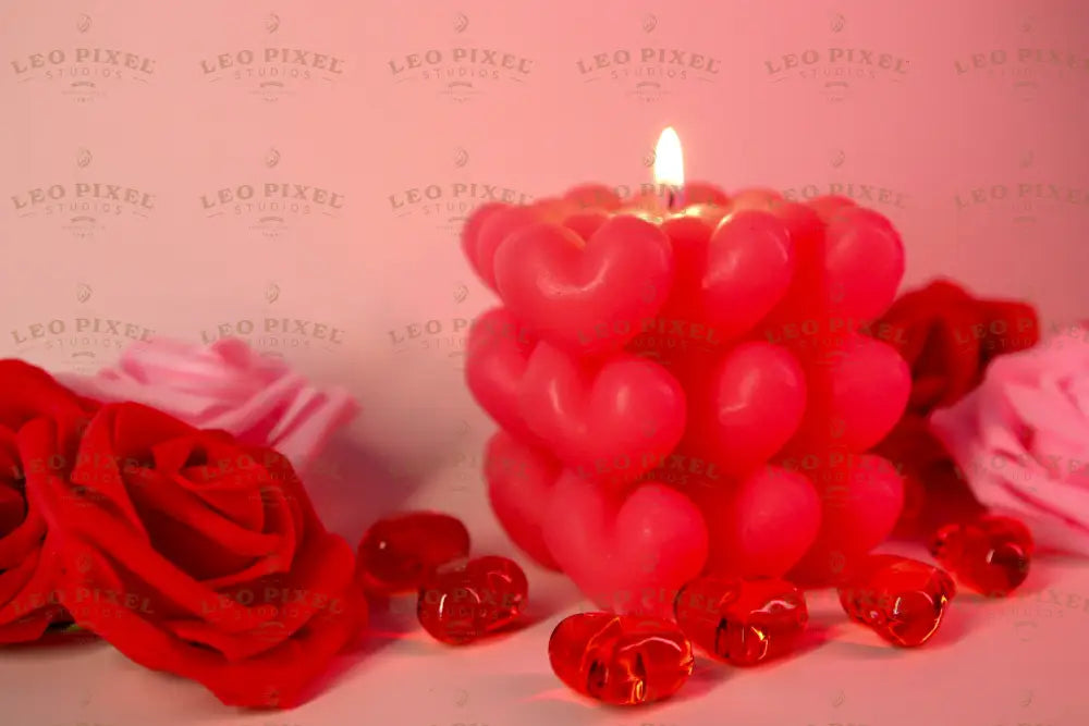 A pink candle made of heart-shaped wax sits in the center with its flame burning. Around it, red and pink artificial roses are arranged on a soft pink surface. Transparent red heart-shaped gems are scattered nearby, reflecting the warm light. The soft background and gentle lighting create a balanced composition, highlighting the candle's texture and details. The close-up angle captures fine elements clearly. Photography.