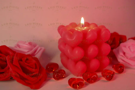 A pink candle made of heart-shaped wax sits in the center with its flame burning. Around it, red and pink artificial roses are arranged on a soft pink surface. Transparent red heart-shaped gems are scattered nearby, reflecting the warm light. The soft background and gentle lighting create a balanced composition, highlighting the candle's texture and details. The close-up angle captures fine elements clearly. Photography.