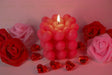 A pink candle made of heart-shaped wax sits in the center with its flame burning. Around it, red and pink artificial roses are arranged on a soft pink surface. Transparent red heart-shaped gems are scattered nearby, reflecting the warm light. The soft background and gentle lighting create a balanced composition, highlighting the candle's texture and details. The close-up angle captures fine elements clearly. Photography.