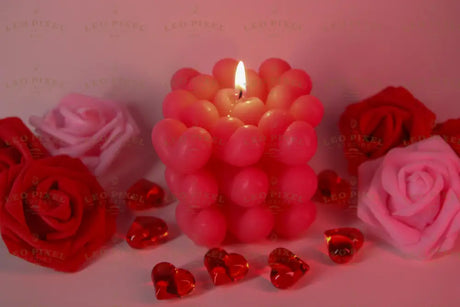 A pink candle made of heart-shaped wax sits in the center with its flame burning. Around it, red and pink artificial roses are arranged on a soft pink surface. Transparent red heart-shaped gems are scattered nearby, reflecting the warm light. The soft background and gentle lighting create a balanced composition, highlighting the candle's texture and details. The close-up angle captures fine elements clearly. Photography.