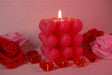 A pink candle made of heart-shaped wax sits in the center with its flame burning. Around it, red and pink artificial roses are arranged on a soft pink surface. Transparent red heart-shaped gems are scattered nearby, reflecting the warm light. The soft background and gentle lighting create a balanced composition, highlighting the candle's texture and details. The close-up angle captures fine elements clearly. Photography.